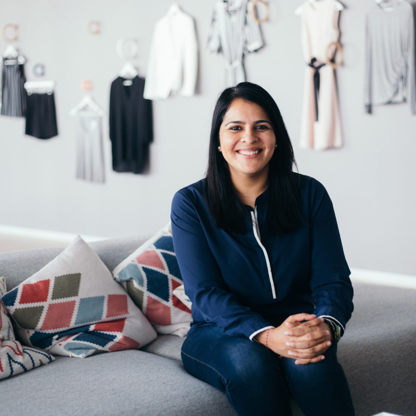 Smiling woman with long black hair wearing a navy blue top, sitting on a gray couch with colorful patterned cushions.