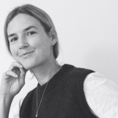 Black and white photo of a woman with light hair smiling gently, resting her chin on her hand, wearing a dark sleeveless sweater over a light shirt and a necklace.