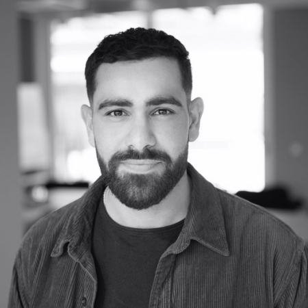 Black and white portrait of a man with short dark hair, beard, and mustache wearing a dark shirt and jacket, smiling slightly against a blurred background.