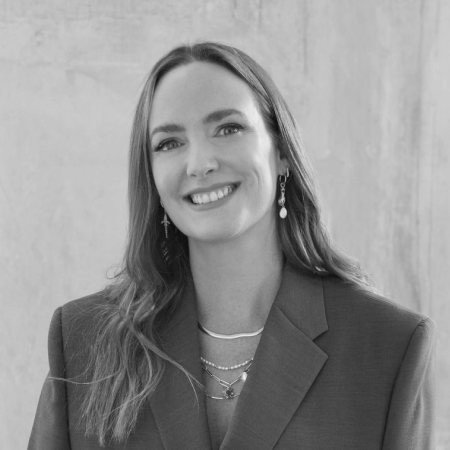 Smiling woman with long hair wearing a blazer and layered necklaces in front of a plain wall.