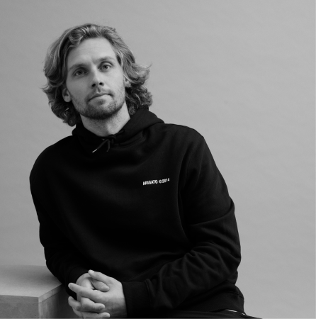 Black and white portrait of a man with medium-length wavy hair wearing a black hoodie sitting and leaning on a surface.