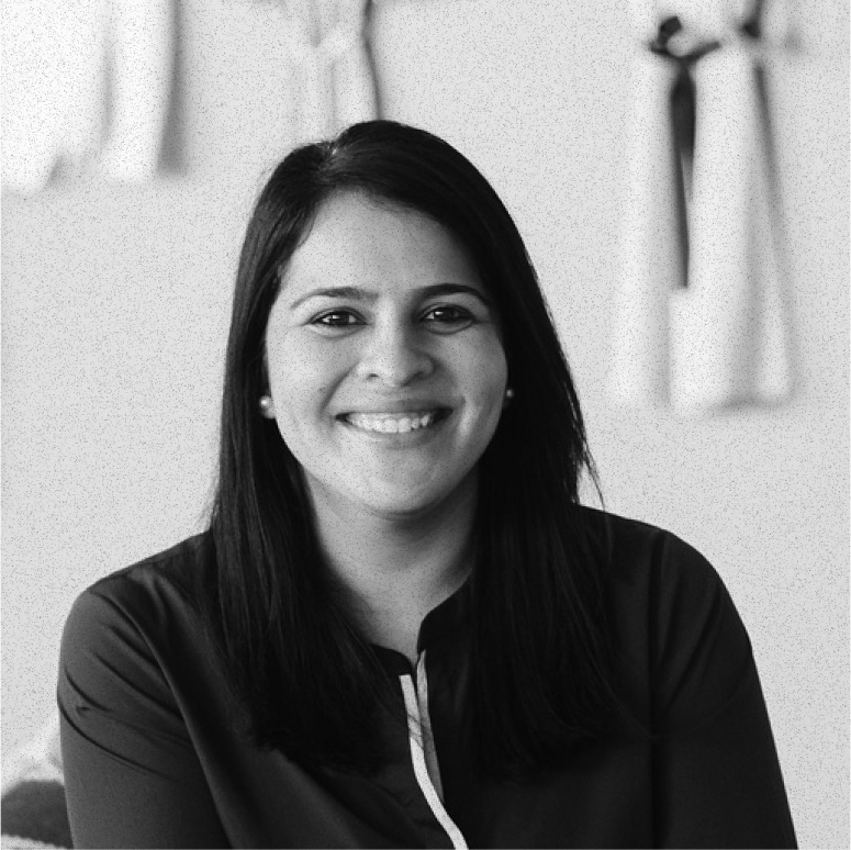 Smiling woman with long dark hair wearing a dark blouse with a white stripe, seated indoors against a blurred background.