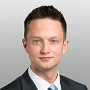 Portrait of a man with short brown hair wearing a suit and tie against a light gray background.